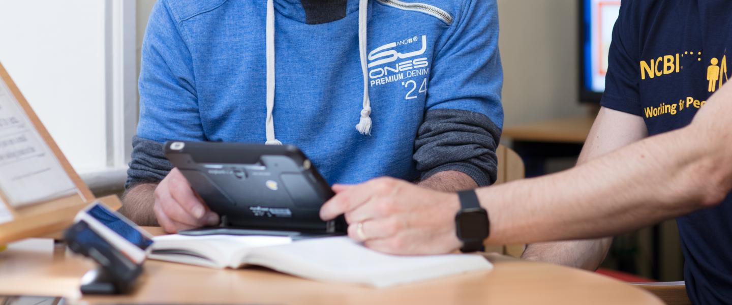 a man in a blue hoodie is using Assistive Technology and an NCBI member is helping him